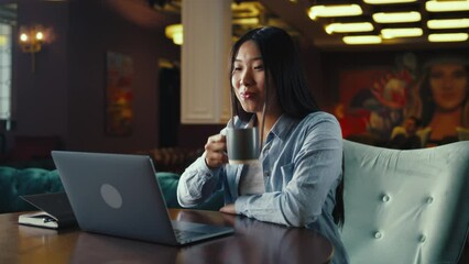 Asian businesswoman looking at screen of laptop, watching business training, working online on startup project, drinking coffee in office cafe. Worker of E-commerce marketing company