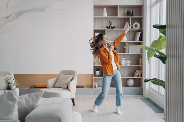 Happy woman dancing to music, using musical mobile phone apps in modern living room at home