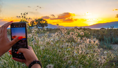 The hand of a girl with a dark bracelet holding a phone and photographing a sunset in the mountains with it