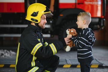 Wall Mural - Portrait of rescued little boy with firefighter man standing near fire truck. Firefighter in fire fighting operation.