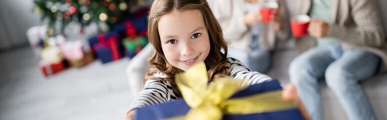 Smiling girl holding blurred christmas present and looking at camera at home, banner