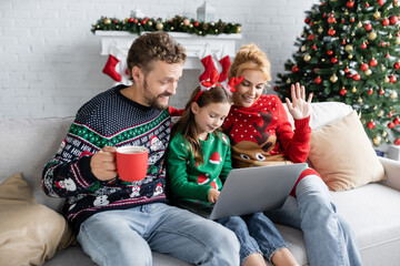 Wall Mural - Smiling family in new year sweaters having video call on laptop at home