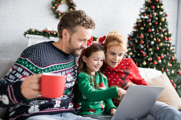 Wall Mural - Smiling girl using laptop with parents in christmas sweaters at home