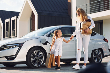 With shopping bags. Young woman with her little daughter is with their electric car outdoors