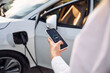 © standret - Smartphone monitoring of charging. Young woman in white clothes is with her electric car at daytime