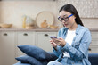 © Liubomir - Young beautiful woman at home sitting in the kitchen smiling and happy, Asian woman using smartphone typing messages and reading news online.