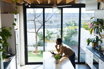 Wall Mural - Happy african american man leaning on countertop in kitchen, using smartphone