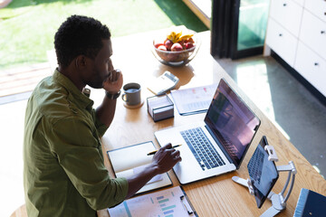 Wall Mural - Happy african american man standing at table in kitchen, using laptop and tablet