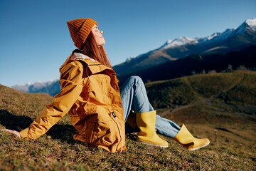 Woman sitting on a hill on the grass rest smile with teeth looking at the mountains in the snow in the autumn in a yellow raincoat and jeans happy sunset hiking trip, freedom lifestyle 