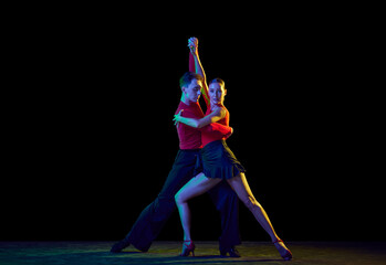 Wall Mural - Full-length portrait of young beautiful man and woman dancing ballroom dance isolated over dark background in neon light. Concept of art, beauty, grace, action, emotions.