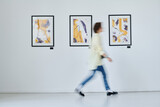 Fototapeta  - Blurred motion of young woman walking along art gallery with modern art on the wall