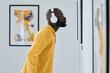 © AnnaStills - African young man in wireless headphones examining painting on the wall at gallery