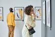 © AnnaStills - Young woman in eyeglasses examining modern art at gallery with other visitors