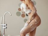 Side view of female sexy body with soap foam indoors in bathroom. Naked woman standing in tub washing body. Close up. Breasts, ass and legs covered in bubbles. Taking a bath