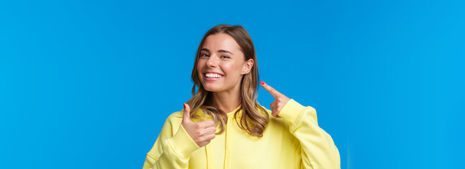 Wall Mural - Cheerful and pleased, satisfied good-looking european female pointing at pierced ear and smiling delighted, show thumbs-up like her new jewelry, standing blue background