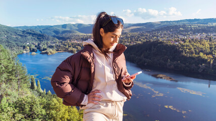 Young woman standing above a lake with stunning view looking her mobile phone 