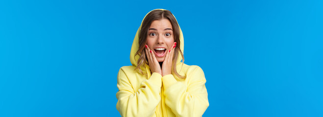 Wall Mural - Close-up portrait of impressed and fascinated blond girl in hood react to amazing great news, touch cheeks gasping and smiling wondered, hear she won prize, standing blue background