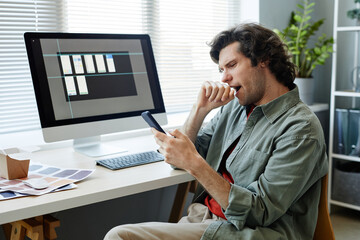 Portrait of Caucasian young man yawning at workplace in office and scrolling social media via phone