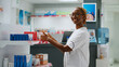 Smiling african american pharmacist checking drugs on shelves in pharmacy, helping clients with medicine, holding pills bottles and boxes of vitamins. Working at drugstore desk. Handheld shot.