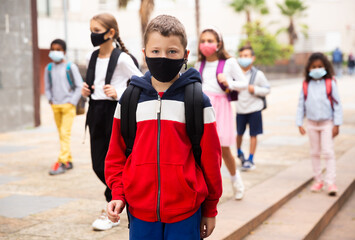 Wall Mural - Portrait of schoolboy in medical mask standing near school, kids on background