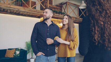 Confident housing agent is showing spacious modern house with beautiful interior to happy young couple wife and husband, people are smiling and talking looking around.