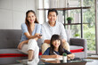 © aekkorn - Children education and home school concept : Young asian father and mother see little daughters' study. Excited smiling small child girl enjoying learning and reading with pleasant dad and mom at home
