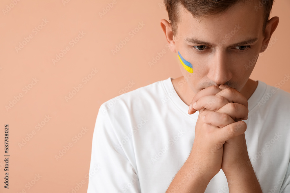 Young man with drawn flag of Ukraine praying on beige background, closeup