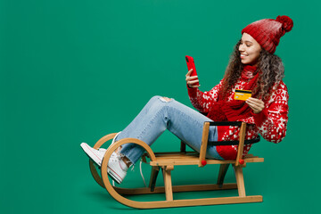 Wall Mural - Full body merry young woman in red sweater hat posing use mobile cell phone credit bank card shopping online order delivery isolated on plain dark green background Happy New Year 2023 holiday concept.