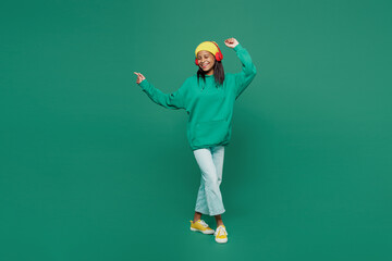 Wall Mural - Full body little kid teen girl of African American ethnicity 13-14 year old in casual hoody hat headphones listen music dance raise up hands isolated on plain dark green background Childhood concept