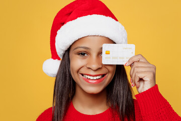 Close up merry little kid teen girl 13-14 year old wear red xmas sweater Santa hat posing hold cover eye with credit bank card isolated on plain yellow background. Happy New Year 2023 holiday concept.