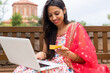 © ADDICTIVE STOCK - Smiling Indian woman sitting on bench with laptop and credit card