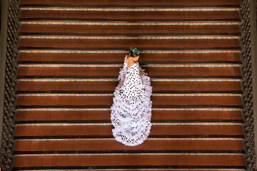 Sticker - Young teenage woman in white dance suit with black polka dots and green carnations in her hair doing flamenco poses on a ladder, top view. Flamenco concept, dance, art, typical Spanish dance.