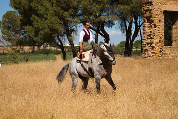 Wall Mural - Young, beautiful Spanish woman on a brown horse in the countryside. The horse raises its front legs. She is doing dressage exercises. Thoroughbred and equine concept.