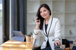 © amnaj - Happy working Asian businesswoman sitting smiling and talking on mobile phone with customers in office.