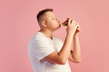 Half-length portrait of cute overweight boy in white t-shirt tasting delicious burger isolated on pink background. Fast food, taste, body positive, emotions