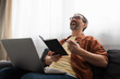 © LIGHTFIELD STUDIOS - excited man in eyeglasses holding pen and notebook while laughing on couch near laptop.