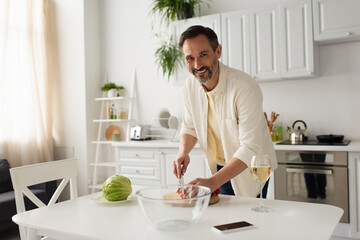 bearded man smiling at camera while cutting cherry tomatoes near fresh lettuce.