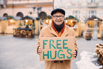 Wall Mural - Happy senior man holding placard with free hugs message at Christmas market and looking at camera.