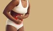 Unknown woman suffering from acute menstrual pain standing on studio beige background. Cropped image of dark skinned woman in underwear holding her hands on red inflamed area of abdomen. Banner.