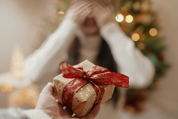 Hand holding christmas gift box on background of happy woman closing eyes waiting for surprise. Merry Christmas and Happy Holidays! Exchanging presents at christmas tree in atmospheric room