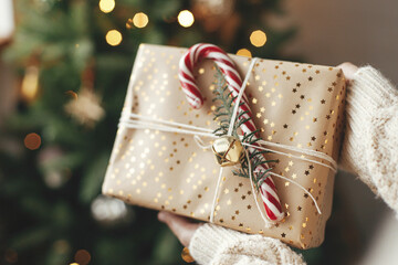 Stylish christmas gift in hands against christmas tree with lights. Merry Christmas and Happy Holidays! Woman in cozy sweater holding wrapped present with candy cane in atmospheric room