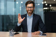 © Liubomir - Video call on online meeting with colleagues, mature boss in business suit looking at web camera and smiling waving to employees, businessman in glasses working inside modern office building.