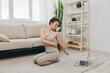 © SHOTPRIME STUDIO - Young Asian woman in front of her laptop doing sports and yoga repeating exercises on video at home, home workout complex for body twisting lifestyle