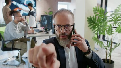 European businessman talks on phone with client and points fingers at camera as if in monitor in company develops 3D games and applications. Against background, studio workers are discussing project