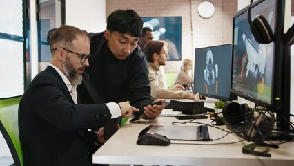 A European man in a business suit discusses a project with an Asian designer showing on a computer monitor in a studio for the development of 3D games and applications