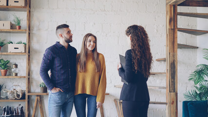 Wall Mural - Attractive young couple is discussing real estate deal with female realtor standing inside beautiful house, looking around and talking. Accommodation and people concept.