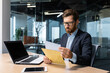 © Liubomir - Serious and focused mature boss working inside modern office building with laptop, businessman in business suit carefully reading letter from bank, upset and disappointed man in glasses and beard.