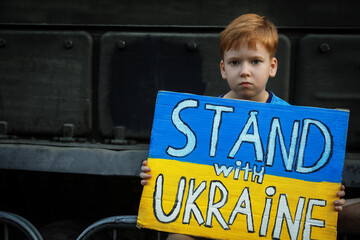 Wall Mural - Sad boy holding poster in colors of national flag and words Stand With Ukraine near broken military tank outdoors. Space for text