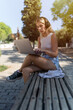 © Juanjo Díaz - A blonde girl with headphones sitting on a bench on a sunny day, using a laptop.