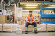 © Maskot - Portrait of smiling young blue-collar worker sitting by hardhat in warehouse